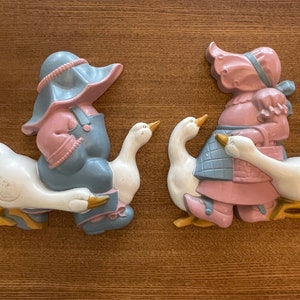May include: Two vintage wall decor pieces featuring a girl and boy riding geese. The girl is wearing a pink dress and blue plaid skirt, while the boy is wearing blue overalls and a pink shirt. Both figures have white geese with gold beaks.