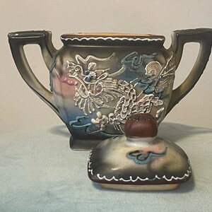 Op de afbeelding: Een theepot van keramiek met een drakenontwerp in tinten bruin, blauw, wit en roze. De theepot heeft een deksel met een knop in de vorm van een paddenstoel. De theepot staat op een lichtblauw oppervlak.