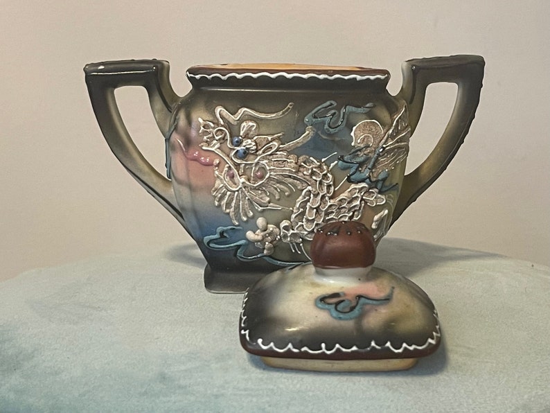 Op de afbeelding: Een theepot van keramiek met een drakenontwerp in tinten bruin, blauw, wit en roze. De theepot heeft een deksel met een knop in de vorm van een paddenstoel. De theepot staat op een lichtblauw oppervlak.