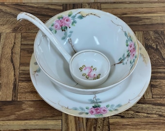 May include: A white porcelain soup bowl with a floral design of pink roses and green leaves. The bowl is nested inside a larger white porcelain plate with a similar floral design. A white porcelain ladle with a gold rim is inside the bowl.