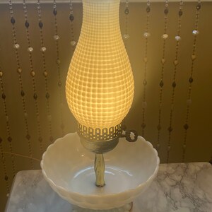 May include: A white milk glass lamp with a bowl base and a textured shade. The lamp is lit and casting a warm glow.