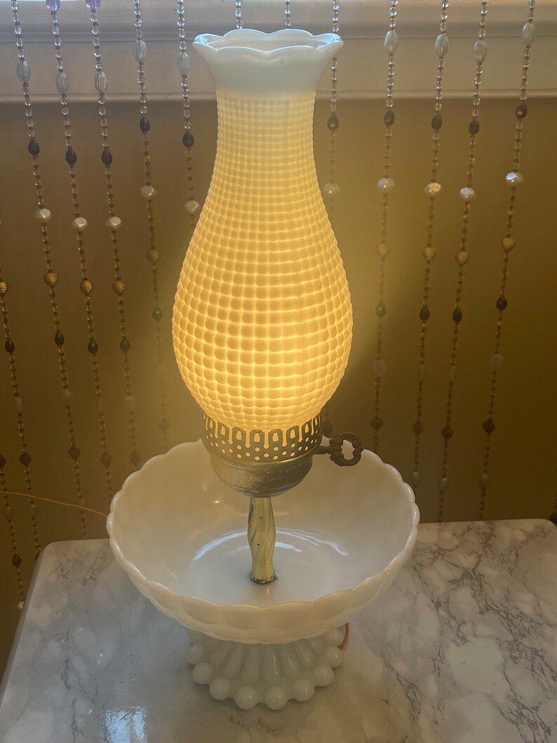May include: A white milk glass lamp with a bowl base and a textured shade. The lamp is lit and casting a warm glow.