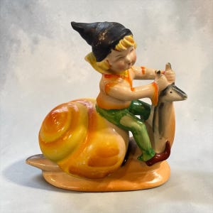 May include: A ceramic figurine of a small figure wearing a black pointed hat, orange shirt, and green pants, riding on the back of a large yellow and orange snail. The figure holds onto the snail's antennae with both hands in a seated position.