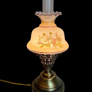 May include: A vintage-style table lamp with a floral design on the shade. The lamp features a brass base and a clear glass chimney. The shade is cream-colored with a red trim and a ruffled edge. The lamp is lit, casting a warm glow.