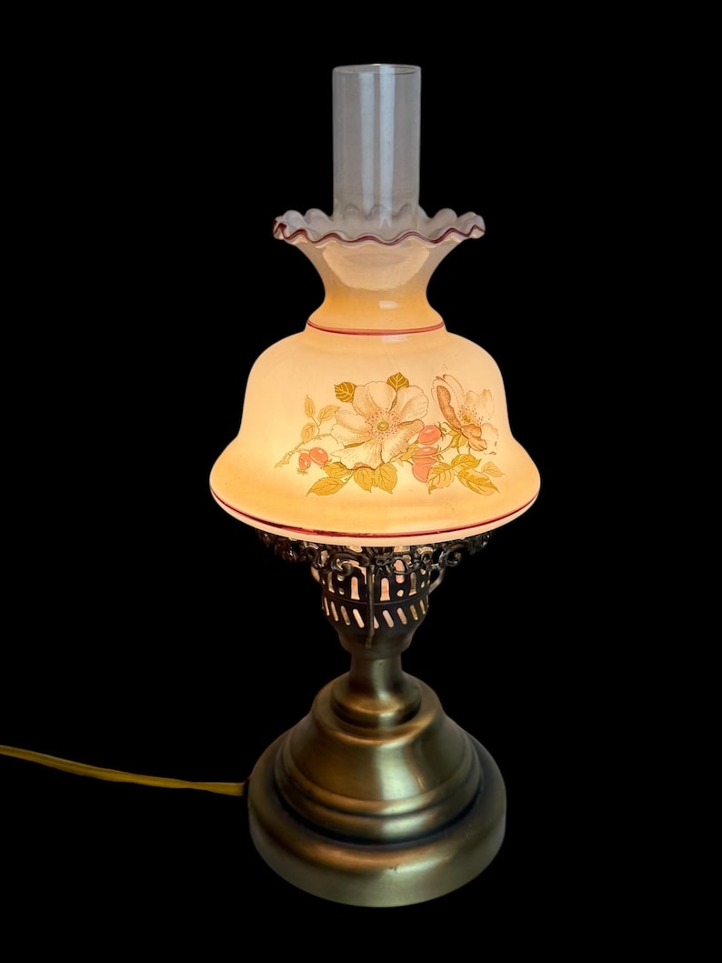 May include: A vintage-style table lamp with a floral design on the shade. The lamp features a brass base and a clear glass chimney. The shade is cream-colored with a red trim and a ruffled edge. The lamp is lit, casting a warm glow.