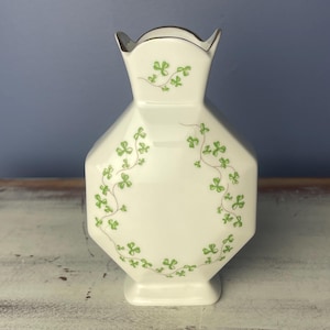 May include: White ceramic vase with a green shamrock design. The vase has a scalloped top and a hexagonal body.