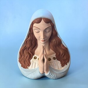 May include: A ceramic bust of a figure with long wavy brown hair and closed eyes, wearing a light blue head covering and a white garment with gold-toned circular accents. The figure is depicted with hands pressed together in a prayer position.