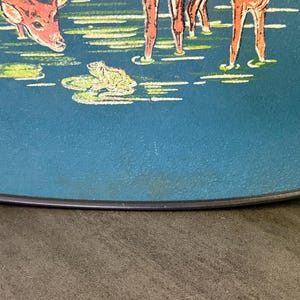 May include: A teal-colored decorative plate with a painted scene of deer in a pond. The deer are brown and white, and the pond has green lily pads and a yellow frog. The plate has a black rim.
