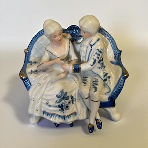 May include: A porcelain figurine depicting a seated couple in 18th-century attire. The figures are dressed in white and blue, with gold accents. They are seated on a blue and white ornate chair. The figurine is approximately 6 inches tall.