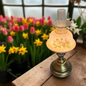 May include: A vintage-style oil lamp with a brass base and a cream-colored glass shade. The shade features a floral design in pink and yellow. The lamp is on a wooden surface, with a blurred background of flowers.