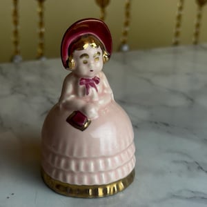 May include: A vintage porcelain figurine of a woman in a pink dress and hat. The figurine has gold accents on the base, hat, and details. The woman is holding a small, red square. The figurine is approximately 4 inches tall.