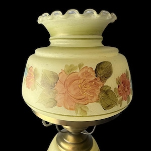 Puede incluir: Una l&aacute;mpara de estilo vintage con una pantalla de cristal amarillo p&aacute;lido. La pantalla presenta flores pintadas a mano en tonos melocot&oacute;n, rosa y verde. La l&aacute;mpara tiene una base de lat&oacute;n y un borde superior con volantes.