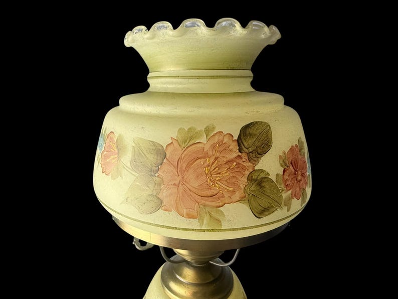 Puede incluir: Una l&aacute;mpara de estilo vintage con una pantalla de cristal amarillo p&aacute;lido. La pantalla presenta flores pintadas a mano en tonos melocot&oacute;n, rosa y verde. La l&aacute;mpara tiene una base de lat&oacute;n y un borde superior con volantes.