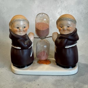May include: A vintage figurine featuring two smiling monk figures flanking a glass hourglass. The monks wear brown robes and hold a wire that supports the hourglass. The hourglass contains pink sand and sits on a white base.
