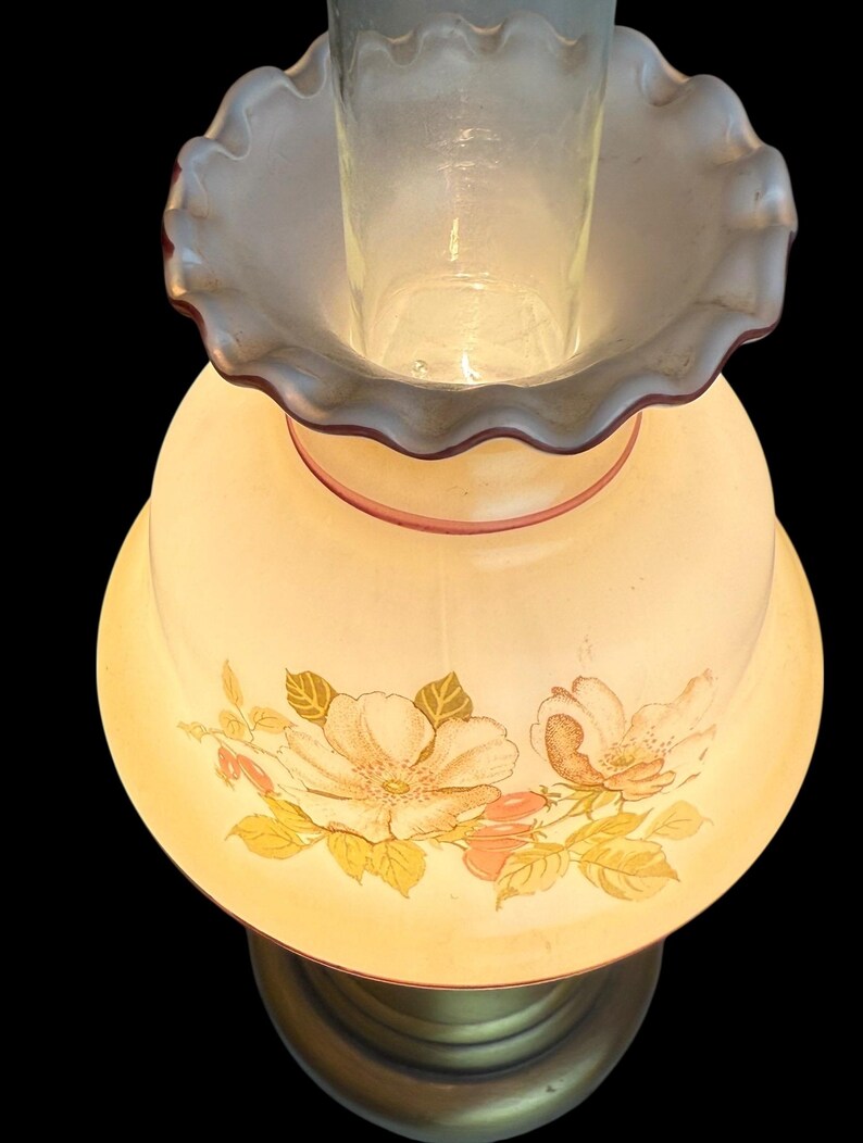 May include: A vintage-style table lamp with a floral design. The lamp features a cream-colored shade adorned with peach and yellow flowers and green leaves. The base is a brass color, and the lamp has a clear glass chimney.