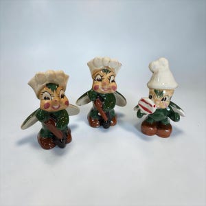 May include: Three ceramic elf figurines with painted features, wearing green outfits, brown shoes, and white hats. Two elves hold brown shovels, while the third holds a round white and red striped object. Each figure has wings and rosy cheeks.