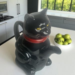 May include: A black ceramic teapot shaped like a cat, with yellow eyes and a red collar. The teapot has a handle and a spout, and is sitting on a white surface. A bowl of green apples is in the background.