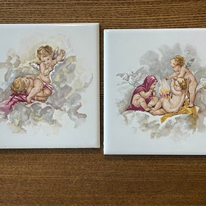 May include: Two vintage ceramic tiles with a white background and a painted image of cherubs. The first tile shows two cherubs in the clouds, one with a red and white robe. The second tile shows three cherubs in the clouds, one with a red robe and two with wings.