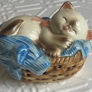May include: A ceramic figurine of a white cat sleeping in a brown wicker basket with a blue blanket. The cat has brown eyes and a pink nose.
