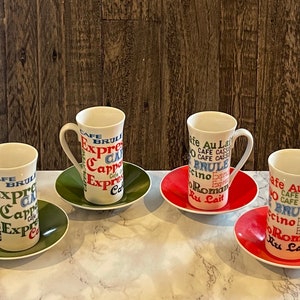 May include: Four white ceramic coffee mugs with colorful text and designs. The mugs are on matching saucers with green, red, and white colors. The text on the mugs includes "Cafe Brule", "Expresso", "Cappuccino", "Cafe Au Lait", and "Romano".