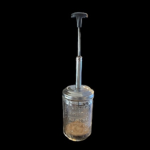 May include: A vintage glass cocktail shaker with a metal lid and handle. The glass jar has measurement markings. The handle is black and the shaker is against a black background.