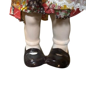 May include: Close-up of a porcelain doll's legs and feet. The doll wears white socks and shiny, black Mary Jane shoes. The doll's dress is visible, featuring a patchwork design with red, yellow, and white floral patterns.