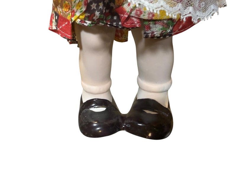 May include: Close-up of a porcelain doll's legs and feet. The doll wears white socks and shiny, black Mary Jane shoes. The doll's dress is visible, featuring a patchwork design with red, yellow, and white floral patterns.