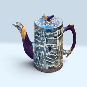 May include: A ceramic teapot featuring a cylindrical body with a black and white marbled pattern, a dark purple handle and spout with metallic silver accents, and a matching lid topped with a small, pointed, dark purple decorative finial.