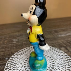 May include: A vintage ceramic figurine of Mickey Mouse standing on a blue base. He is wearing a yellow shirt, blue pants, and black shoes. His ears are black and his face is painted with a friendly expression.