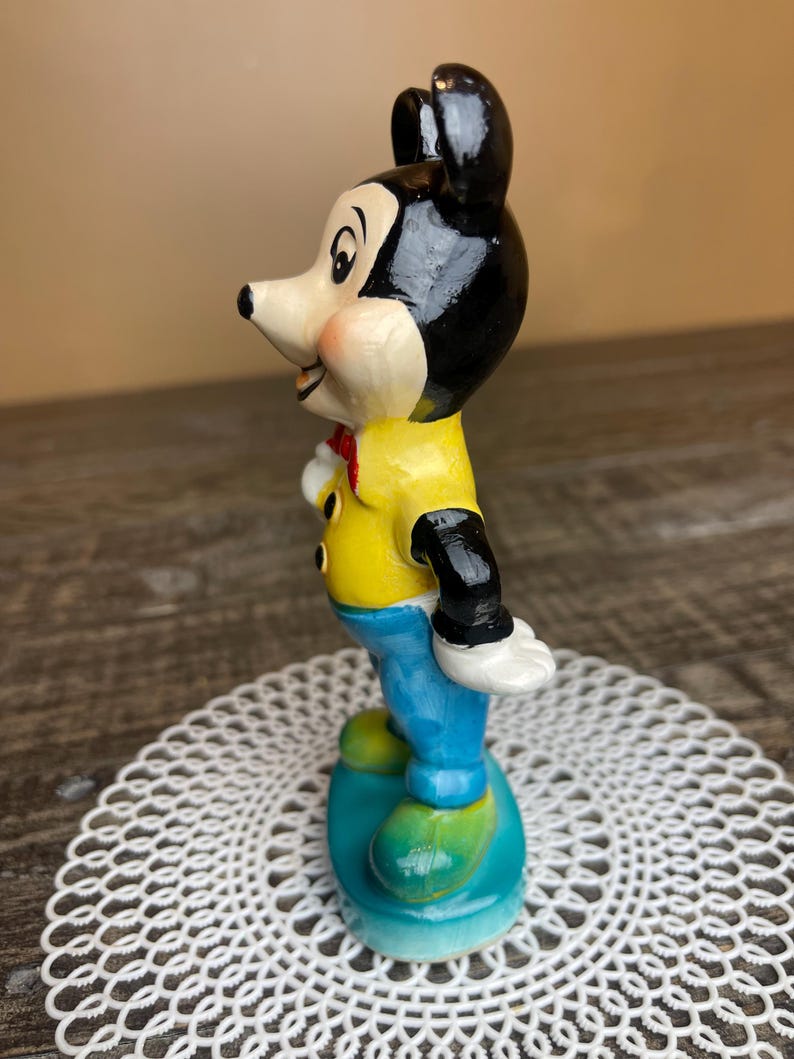 May include: A vintage ceramic figurine of Mickey Mouse standing on a blue base. He is wearing a yellow shirt, blue pants, and black shoes. His ears are black and his face is painted with a friendly expression.
