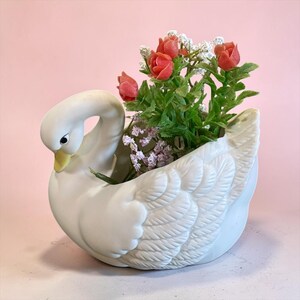 May include: A white ceramic swan-shaped planter featuring a curved neck and detailed wing textures. The hollow body contains a bouquet of artificial flowers with small red rosebuds and delicate white filler blossoms, creating a decorative display.
