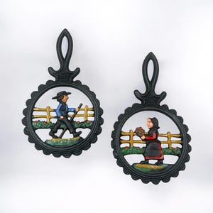 May include: Two black cast iron trivets with a scalloped edge and a handle. Each trivet features a painted scene of people in a field with a fence. One shows a person walking with a stick, the other a person holding flowers.