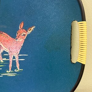 May include: Round serving tray with a dark blue surface and a black rim. The tray features a pink deer illustration and a woven handle. The deer is standing in a grassy area. The tray is a decorative item.