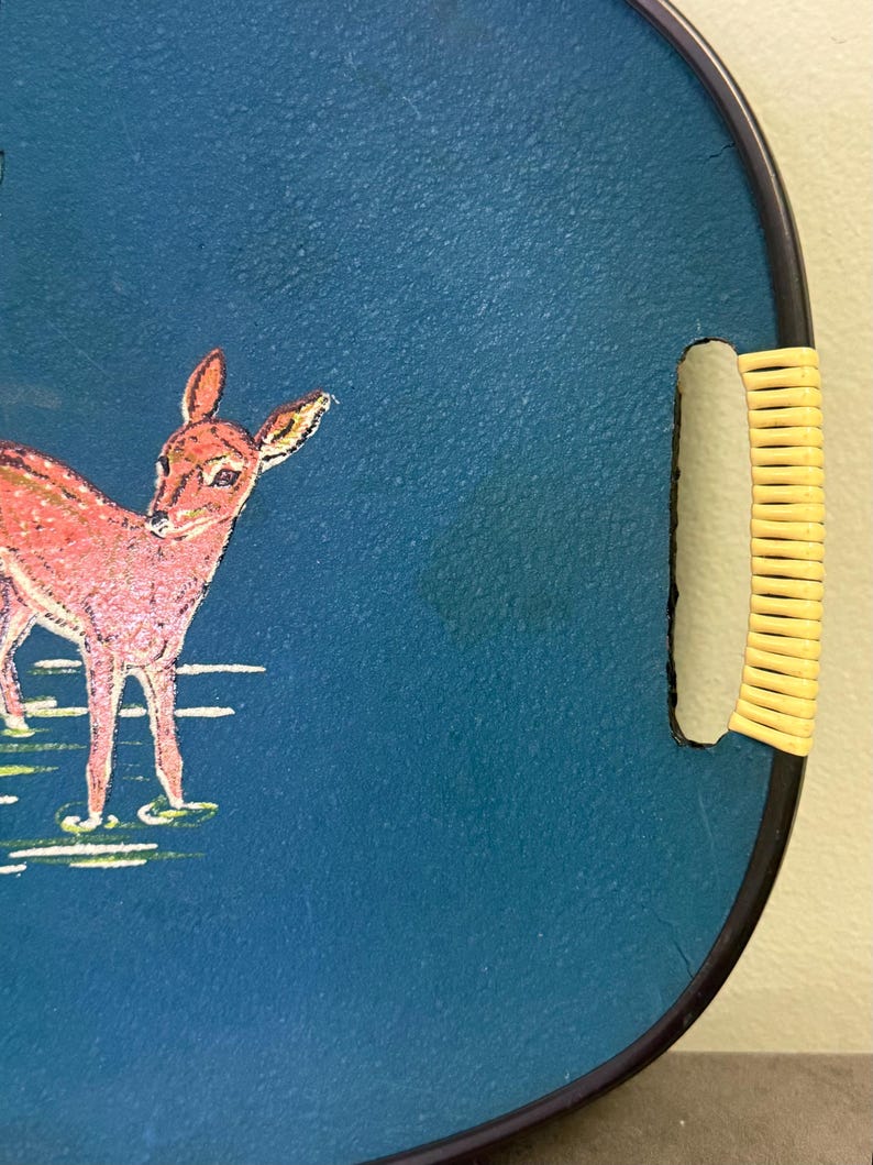 May include: Round serving tray with a dark blue surface and a black rim. The tray features a pink deer illustration and a woven handle. The deer is standing in a grassy area. The tray is a decorative item.