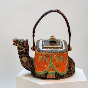 May include: A ceramic teapot shaped like a camel in a seated position. The camel is dark brown with gold and floral accents. It features a bright orange saddle blanket with gold geometric patterns and a woven brown handle attached to the top.