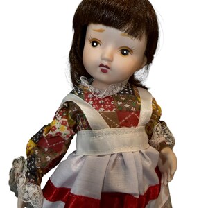 May include: A porcelain doll with brown hair and a detailed face. The doll wears a patchwork print dress with a white apron and red trim. The doll's sleeves have lace cuffs.
