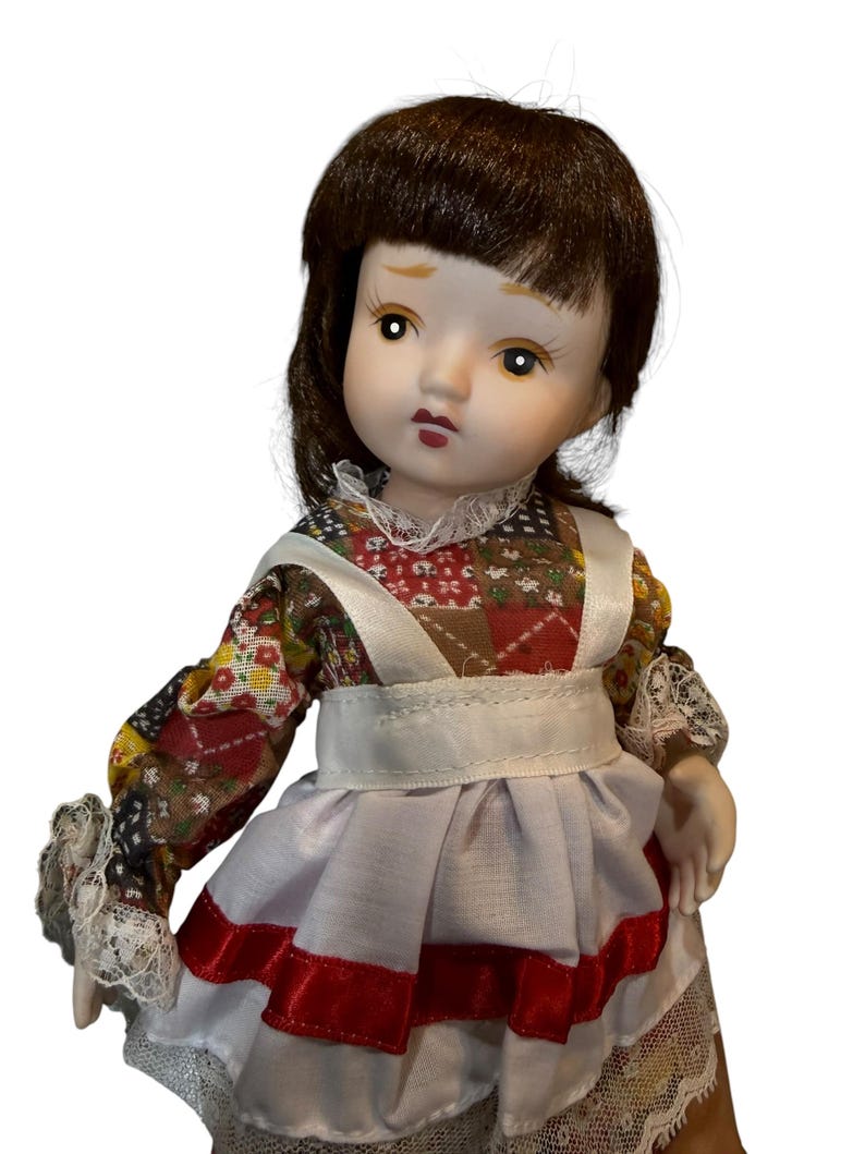 May include: A porcelain doll with brown hair and a detailed face. The doll wears a patchwork print dress with a white apron and red trim. The doll's sleeves have lace cuffs.