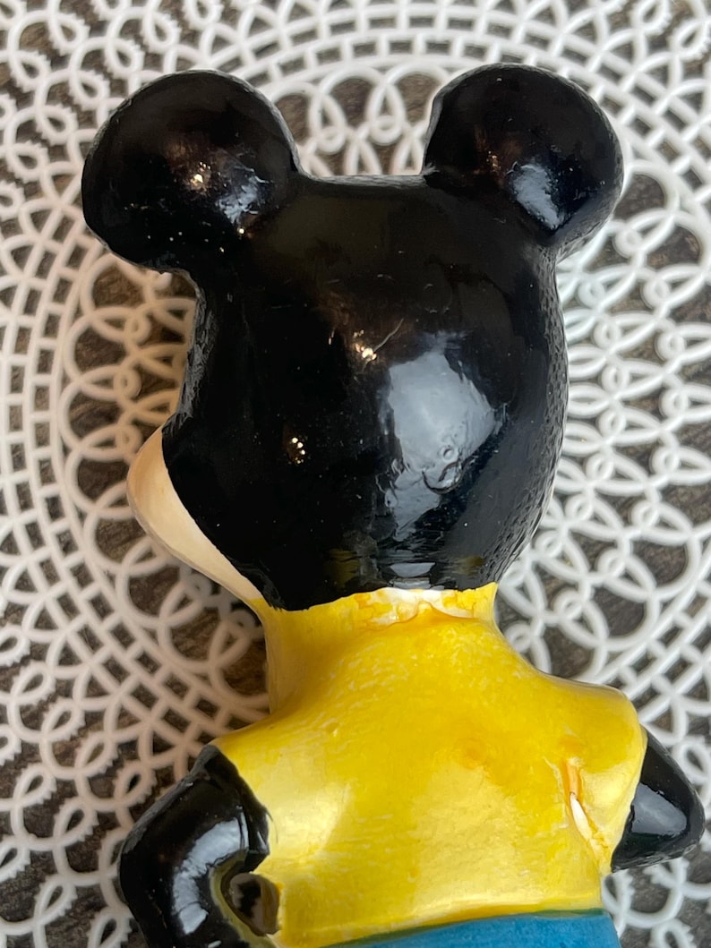 May include: A vintage ceramic figurine of Mickey Mouse. The figurine is painted black, yellow, and blue. Mickey is wearing a yellow shirt and blue pants.