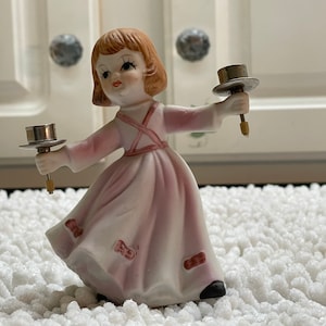 May include: A porcelain figurine of a girl in a pink dress holding two candle holders. The candle holders are silver and have a round base.