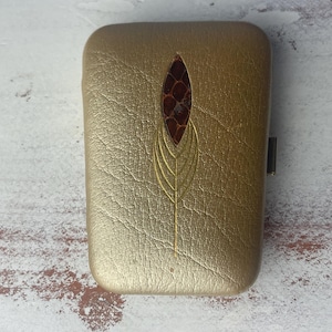 May include: A rectangular, gold-colored case with a textured surface. The case features a central design of a brown, textured oval shape, framed by a gold leaf-like pattern. A small, black clasp is visible on the side.