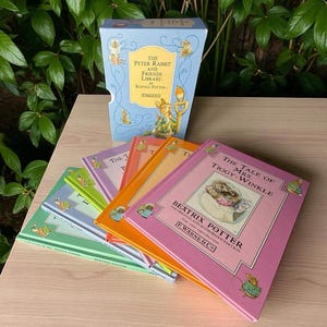 May include: A collection of Beatrix Potter books, including "The Tale of Mrs. Tiggy-Winkle," in a light blue box set. The books have colorful covers in shades of green, purple, orange, and pink. The box set is titled "The Peter Rabbit and Friends Library."