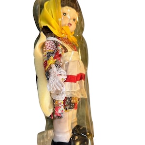 May include: A vintage doll in a clear plastic package. The doll wears a yellow headscarf, a floral patterned top, and a white apron with red and white trim. The doll has black shoes and is in a standing position.