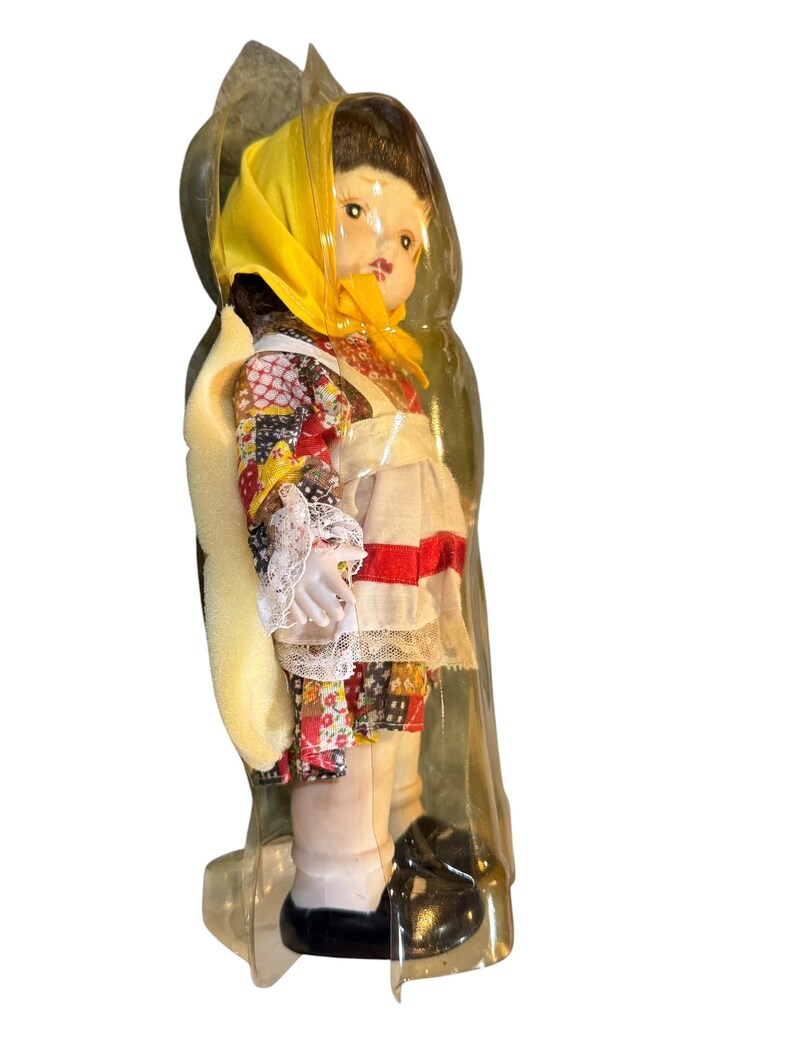 May include: A vintage doll in a clear plastic package. The doll wears a yellow headscarf, a floral patterned top, and a white apron with red and white trim. The doll has black shoes and is in a standing position.