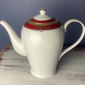 May include: A white porcelain teapot with a red and gold trim. The teapot has a classic design with a rounded body and a spout on one side. The handle is attached to the top of the teapot.