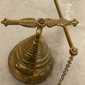 May include: A brass bell with a decorative design and a chain. The bell is attached to a wall mount with a swivel arm.