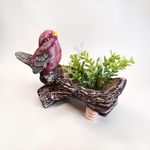 May include: A ceramic bird planter in shades of burgundy, brown, and white. The bird is perched on a faux log, with a small planter area filled with green and white artificial plants. The planter has a textured, rustic appearance.