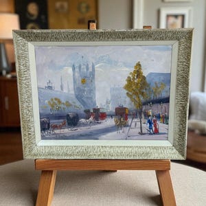May include: An oil painting in a distressed, light green frame, depicting a European street scene with horse-drawn carriages, buildings, and figures. The artwork is displayed on a wooden easel.