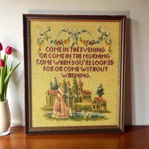 May include: Framed vintage needlepoint artwork with a dark brown wooden frame. The artwork features a yellow background with a quote in burgundy text and a detailed scene of a building and figures. The quote reads: "COME IN THE EVENING OR COME IN THE MORNING COME WHEN YOU'RE LOOKED FOR OR COME WITHOUT WARNING."