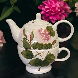 May include: White ceramic teapot and teacup set with a floral rose design. The teapot has a lid and spout, and the teacup is stacked underneath. The set is decorated with pink roses, green leaves, and brown stems.