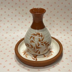 May include: A ceramic vase with a narrow neck and a bulbous body, featuring a glossy white glaze with brown painted leaf patterns and a solid brown rim. The vase sits centered on a matching round ceramic plate with a brown edge and white center.